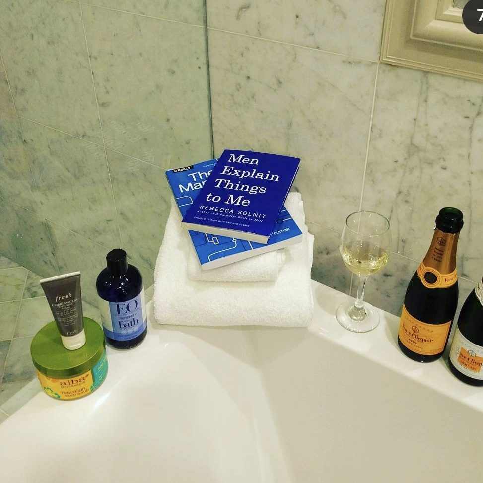An assortment of items sitting on the edge of a bathtub: two demi bottles of champagne, two books, a couple of towels, and toiletries for the bath.